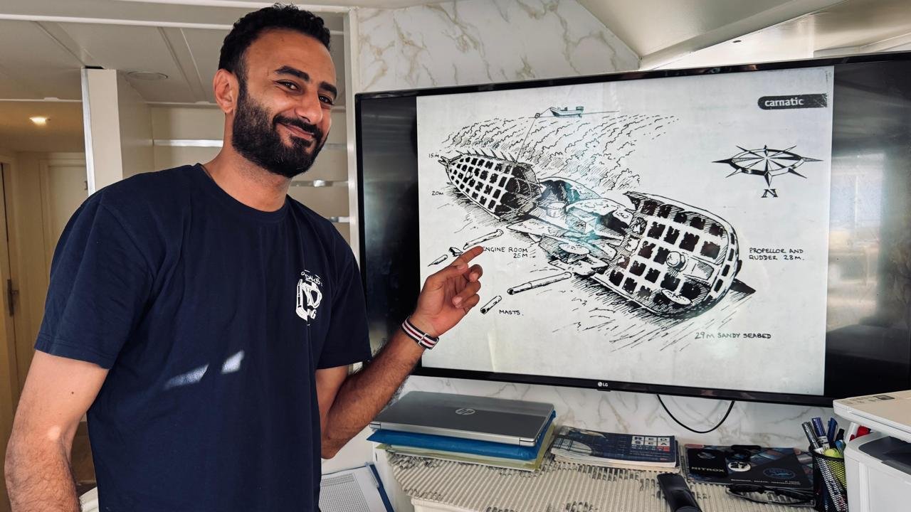 Mahmoud on board, pointing at a drawing of a wreck on the screen during a dive briefing.