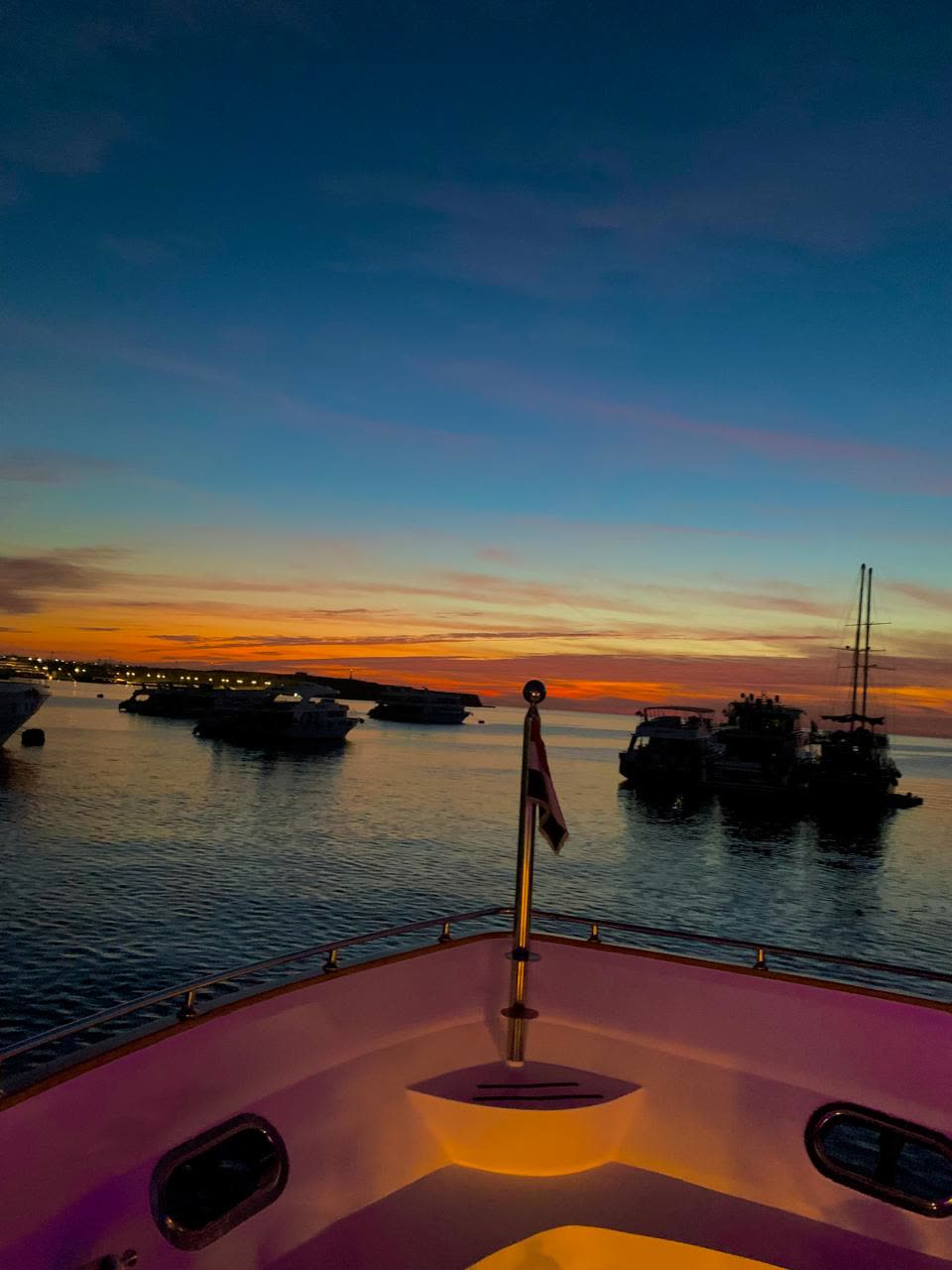 Boats at anchor at dusk, seen over the bow, an orange band of light on the horizon.