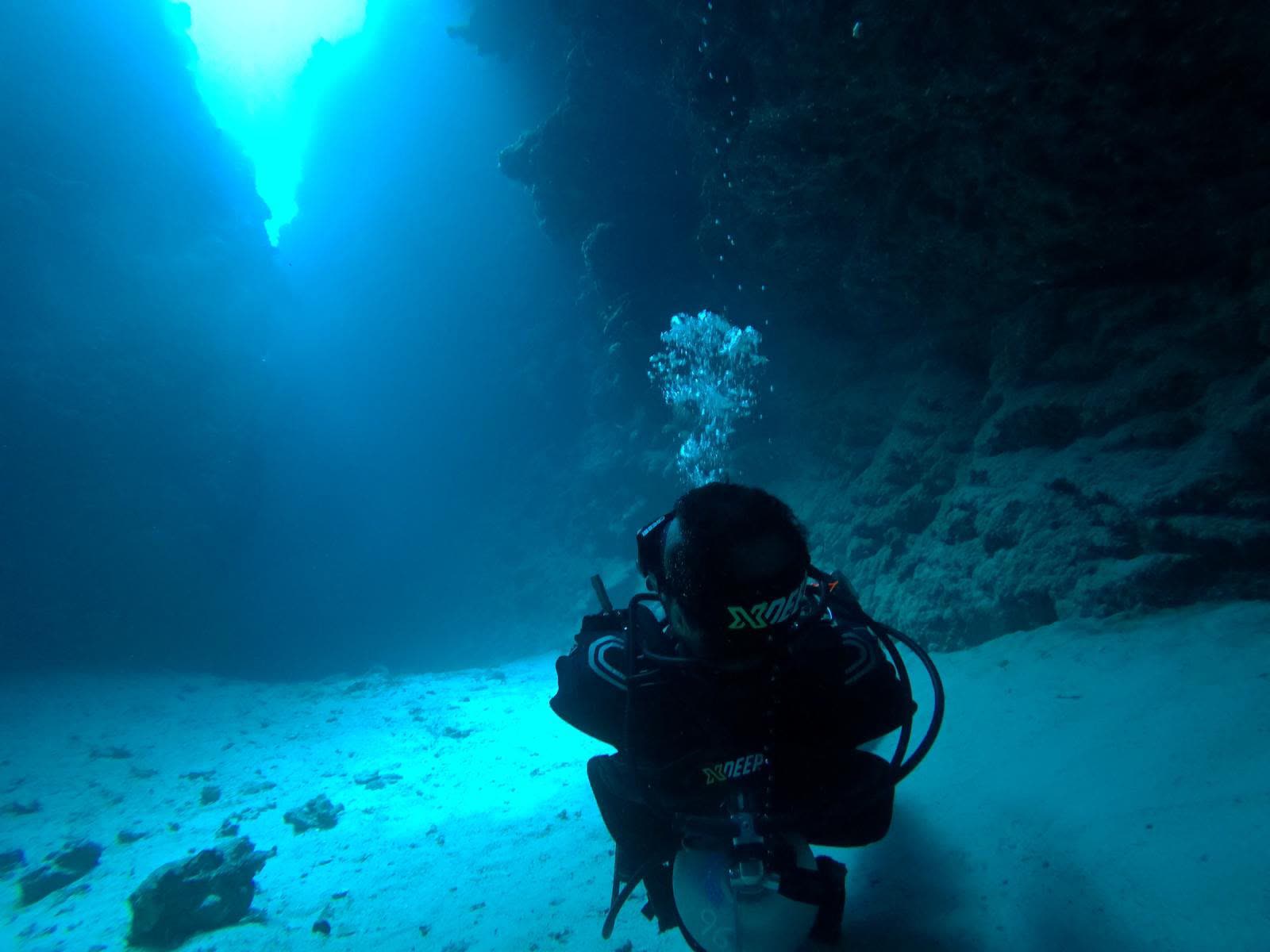 A diver kneeling on the sand inside a canyon, light falling through the opening above.