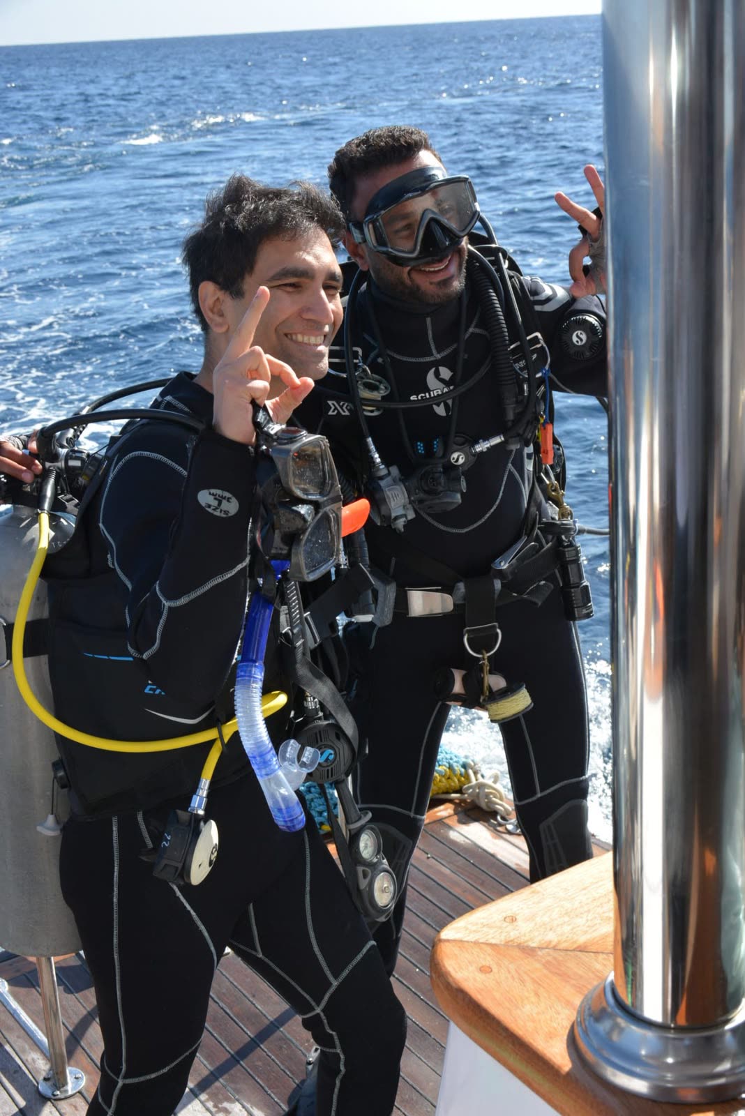 Two divers in full kit on the boat, one waving at the camera.