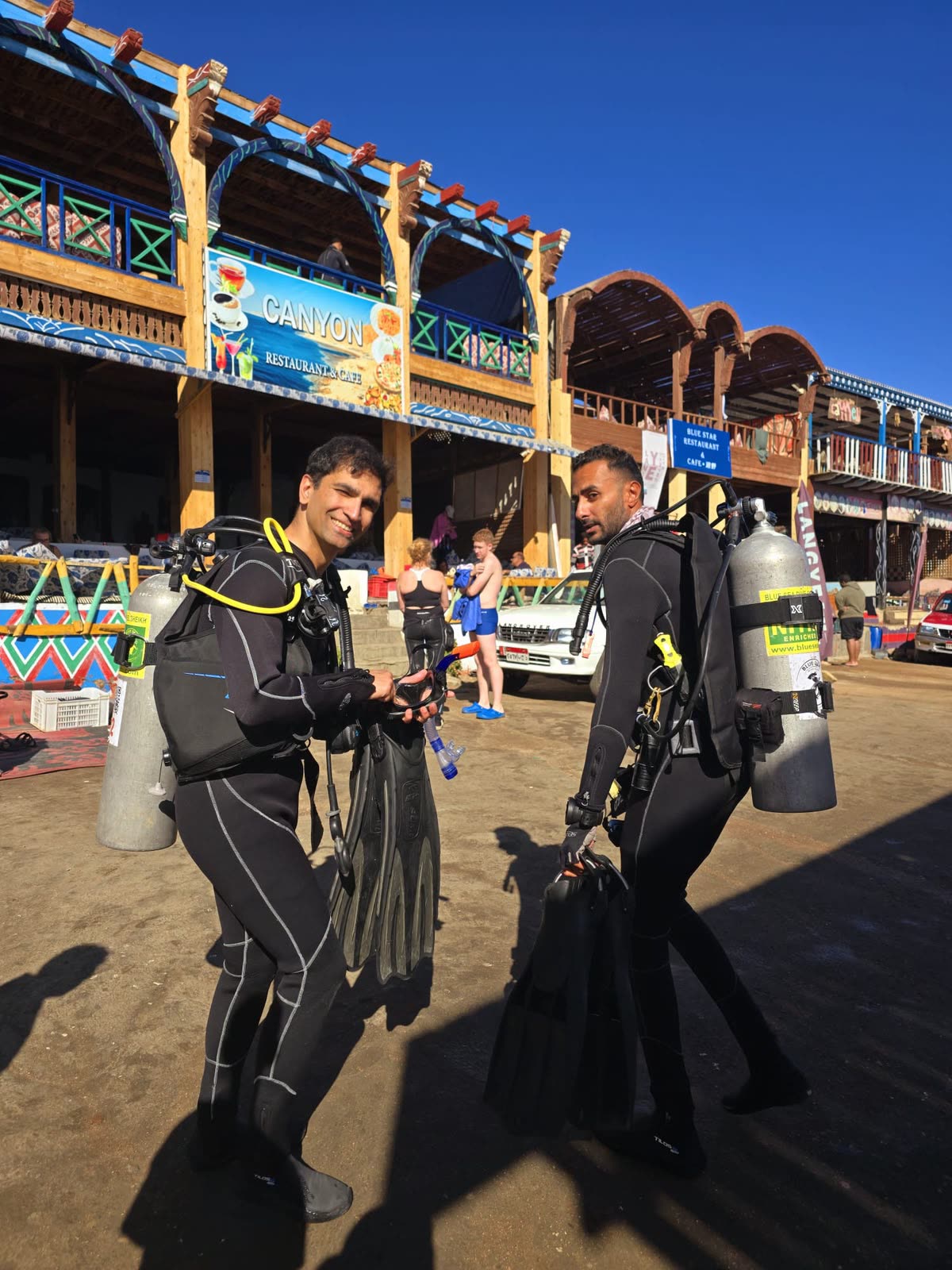 Two divers carrying fins along the Dahab seafront, cafes behind them.