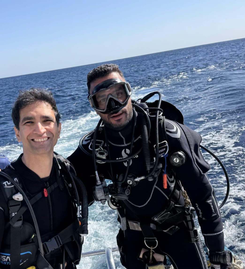 Two divers in wetsuits smiling on the boat, the wake behind them.