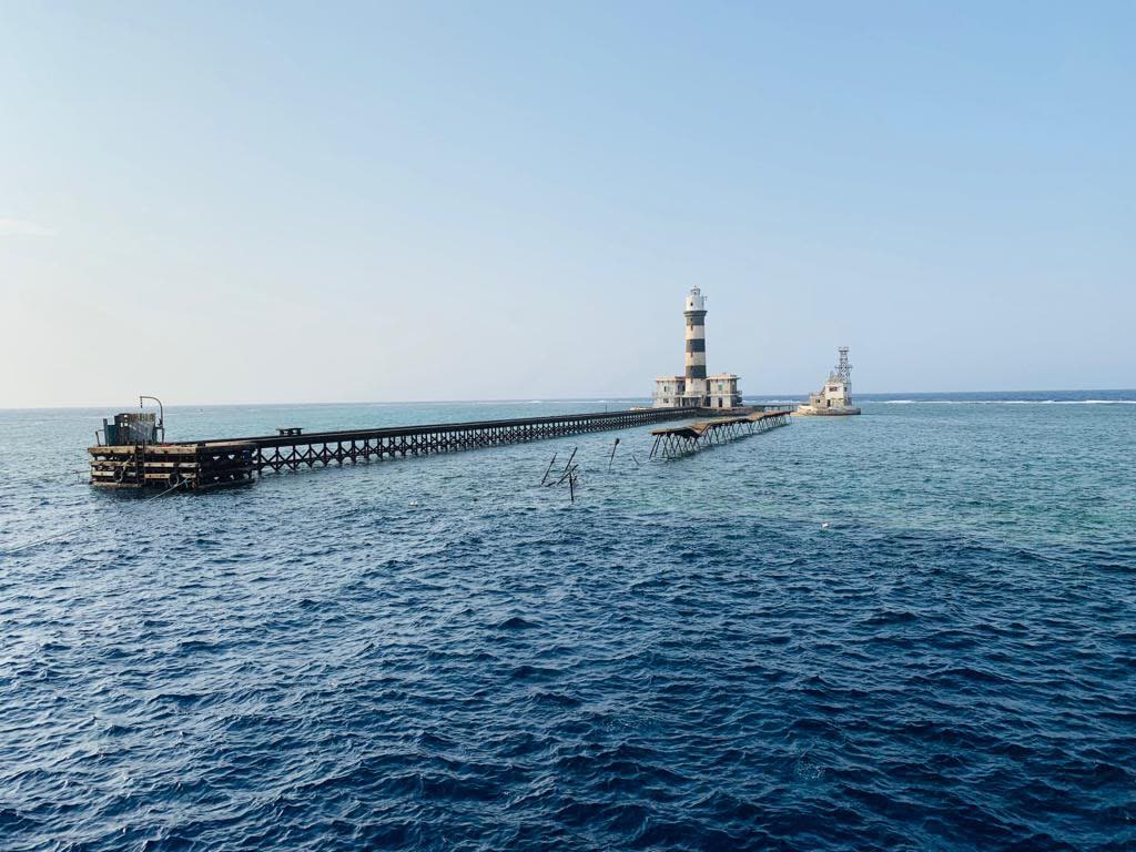 The lighthouse and long jetty on an offshore reef, flat blue sea around it.