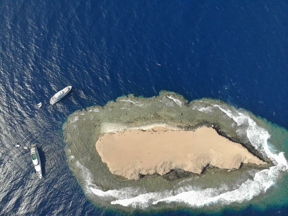 A small sandy island ringed by reef and surf, seen from above, two boats moored nearby.