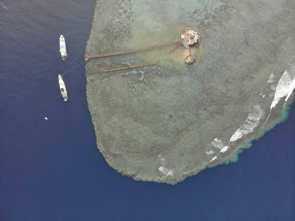 The tip of an offshore reef from above, a small lighthouse and mooring lines running out to boats.