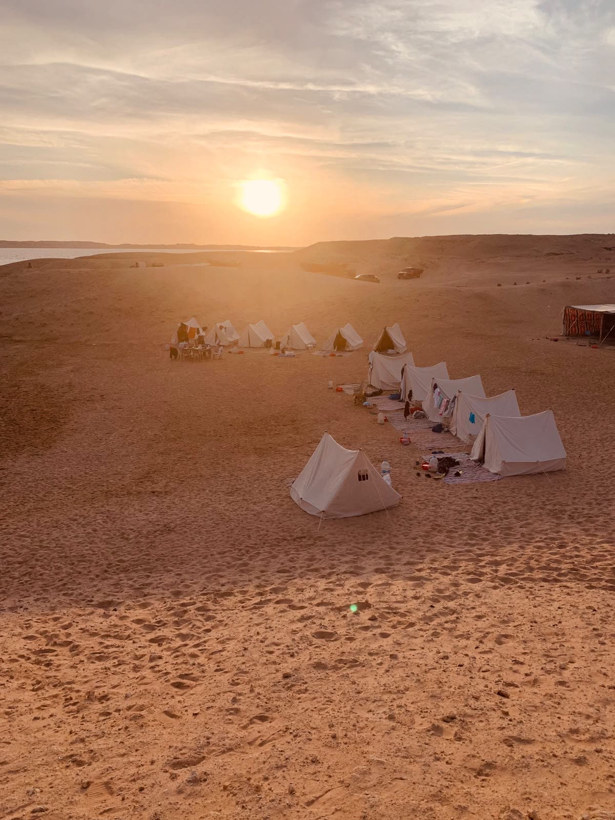 The sun setting behind a scattered camp of white tents on open sand.