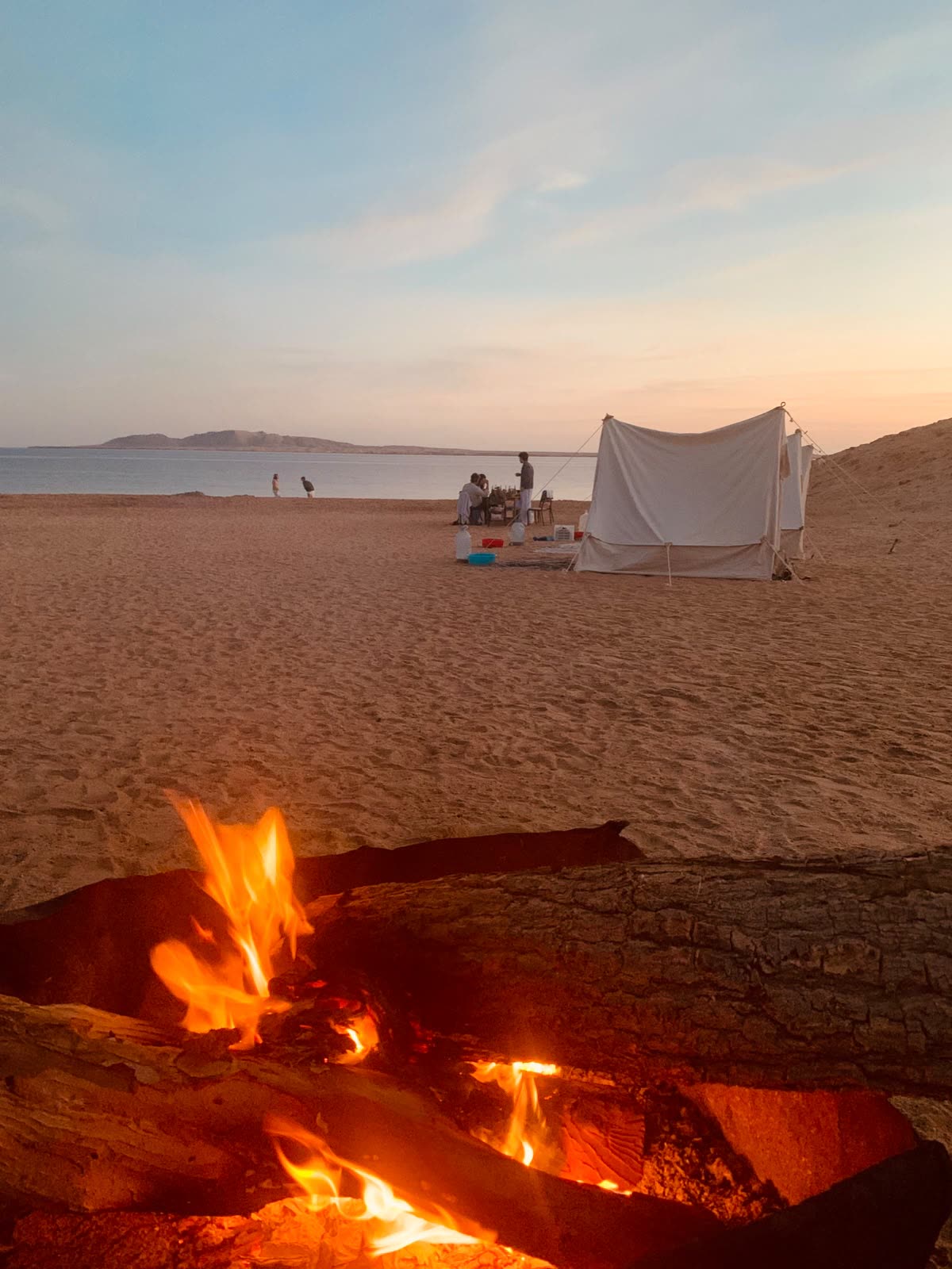 A campfire burning on the sand next to a white tent, people sitting by the water beyond.