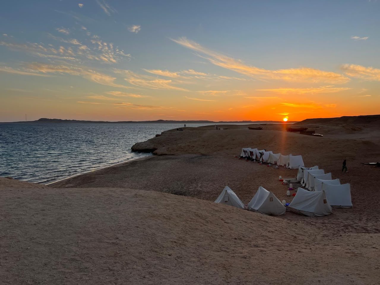 White tents in a curve on the sand above the bay as the sun sets over the sea.