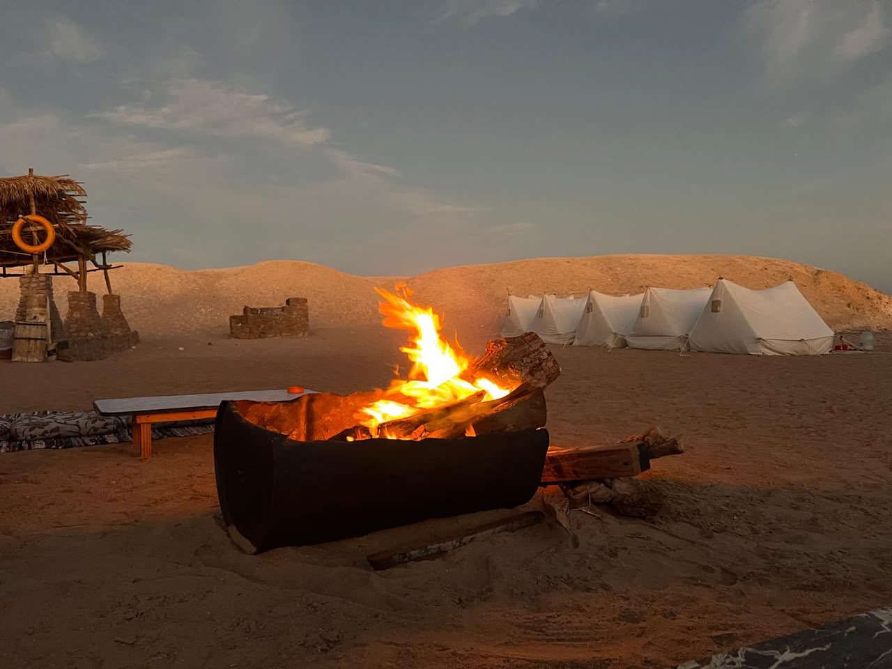 A campfire burning in a metal pit on the sand, a row of white tents behind it at dusk.