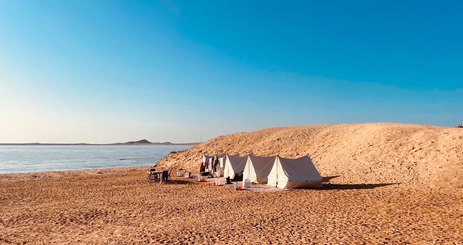 White tents at the foot of a sand dune by the sea, under a clear blue sky.