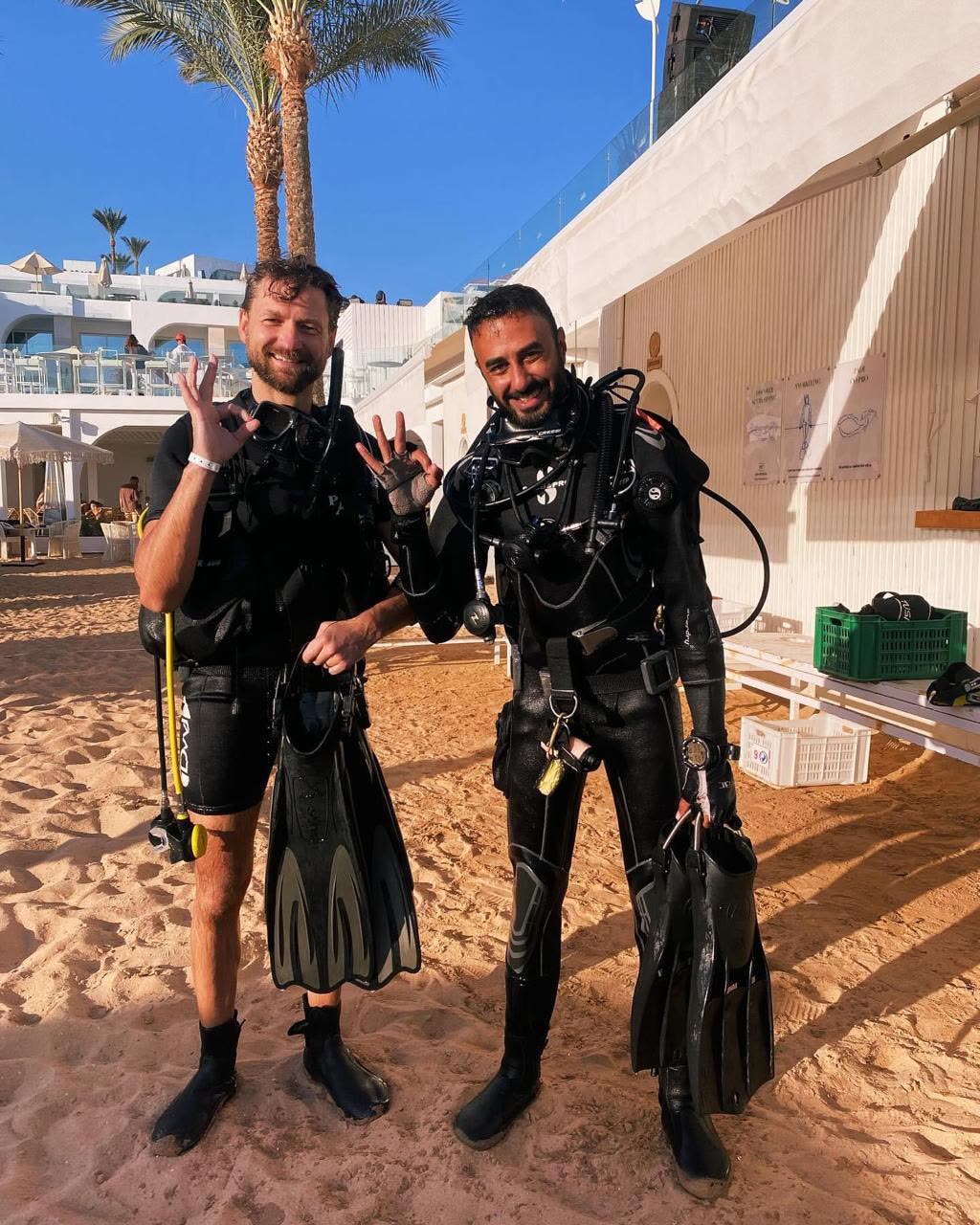 Two divers standing on the sand in wetsuits and BCDs, fins in hand, by the dive centre.