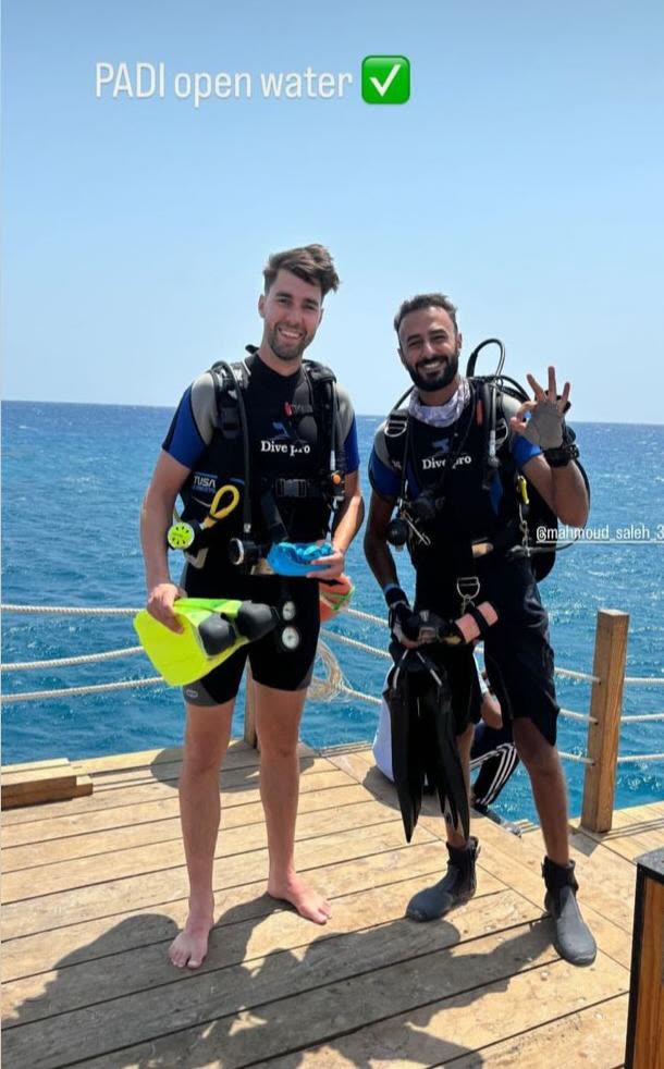 Two divers in full kit on a wooden jetty, the sea behind them, giving the OK sign.