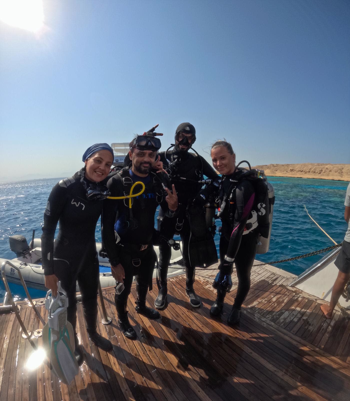 Four divers kitted up on the boat’s dive platform, grinning at the camera, reef and desert coast behind them.