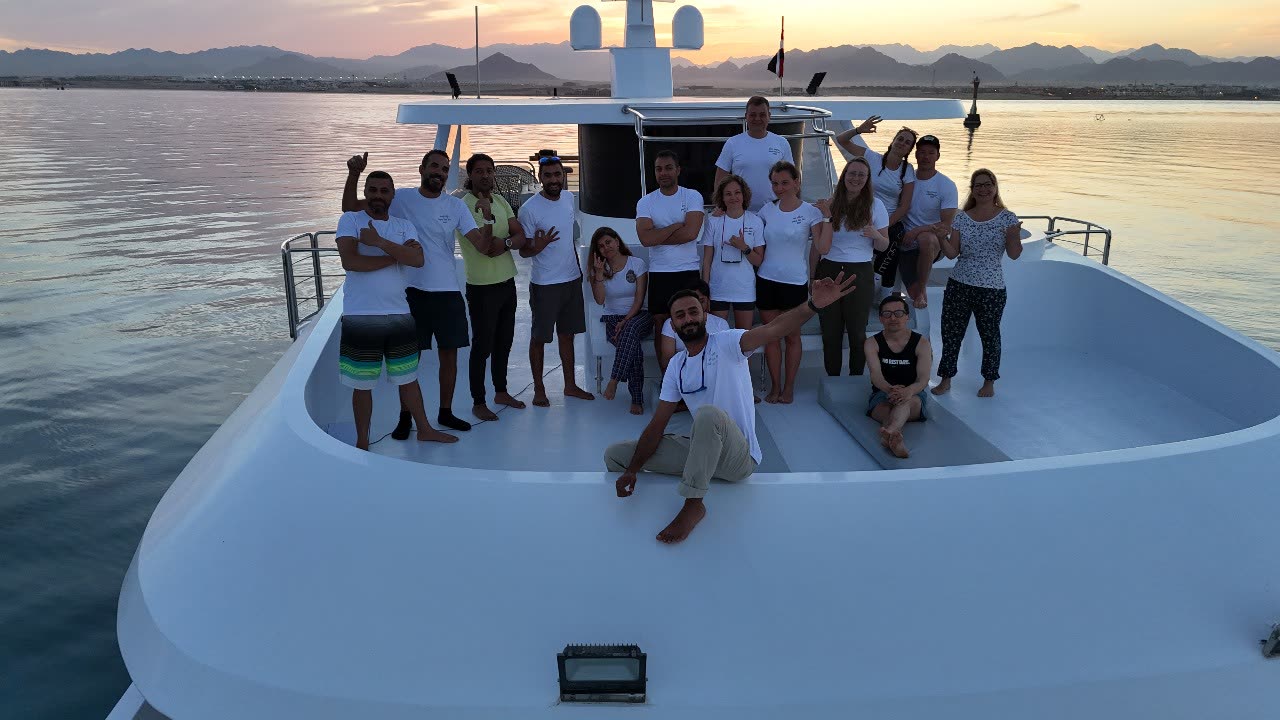 A large group in matching white t-shirts gathered on the bow at the end of the day, waving at the camera, bare desert hills behind.