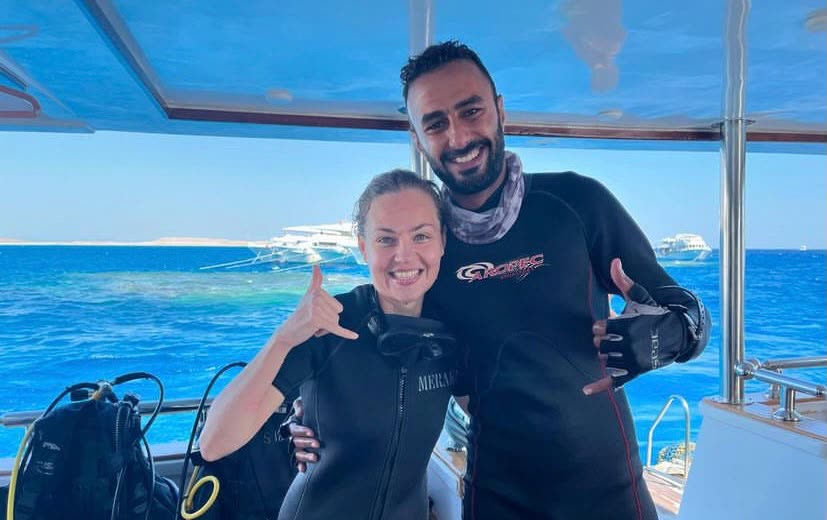 Two divers in wetsuits on the boat, one giving a shaka.