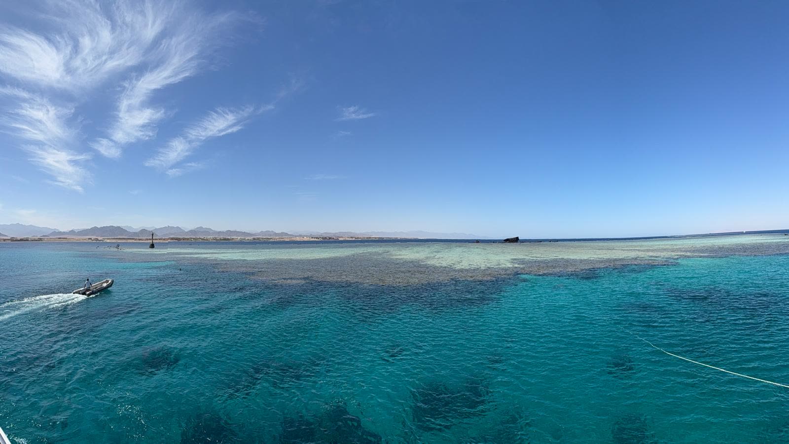 Shallow reef showing pale green through clear water, seen from the deck, with the desert coast low on the horizon.