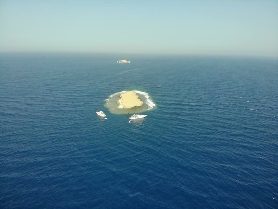 A small sand island alone in open blue water, seen from high above, boats moored beside it.