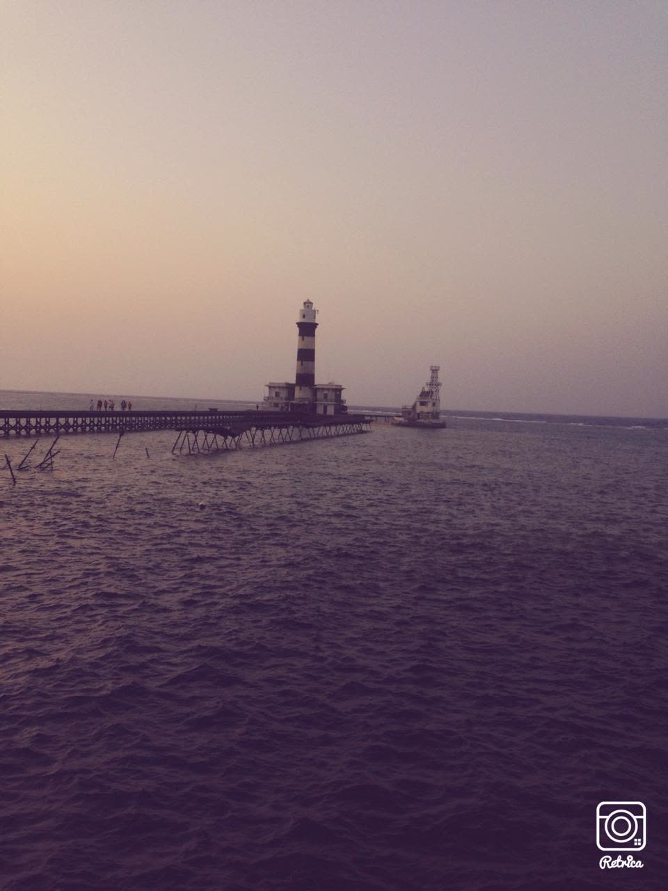 A striped lighthouse on a reef at dusk, a long jetty running out to it across calm water.
