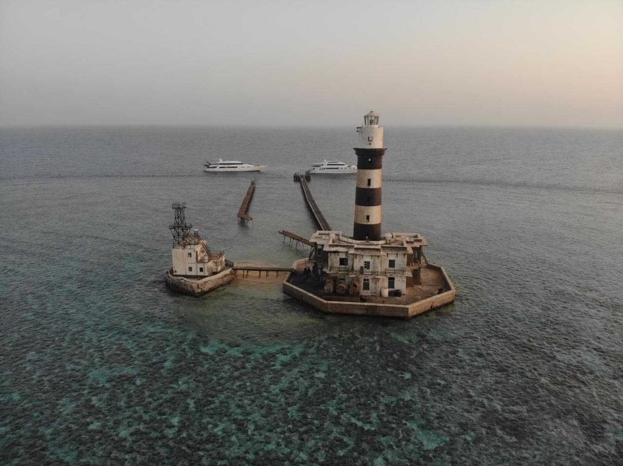 A striped lighthouse and its stone buildings on an offshore reef, liveaboards moored in the distance.