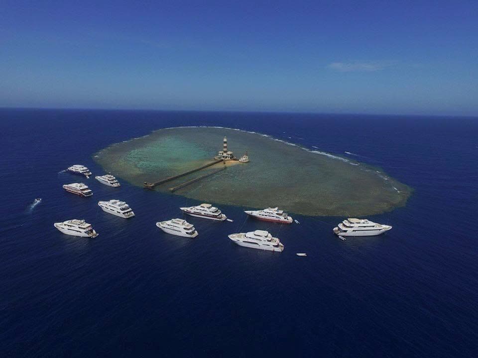 An oval reef from the air with a lighthouse on it, ringed by white liveaboards at their moorings.