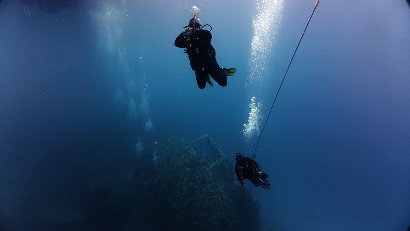 Divers descending a line toward the dark shape of a wreck on the bottom.