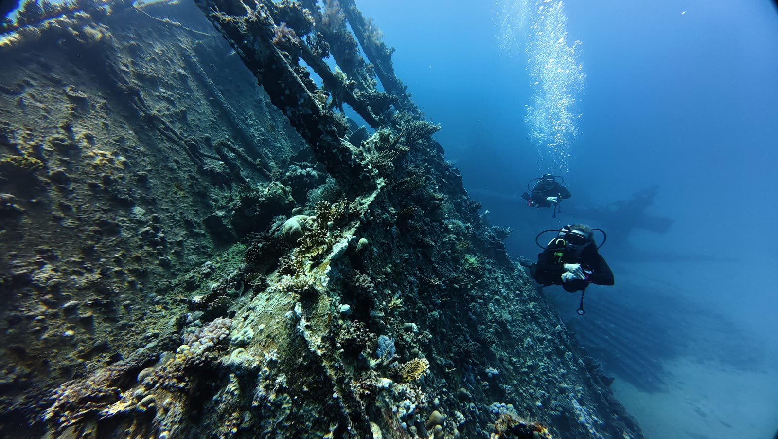 A diver alongside the coral covered hull of a wreck, a second diver further along.