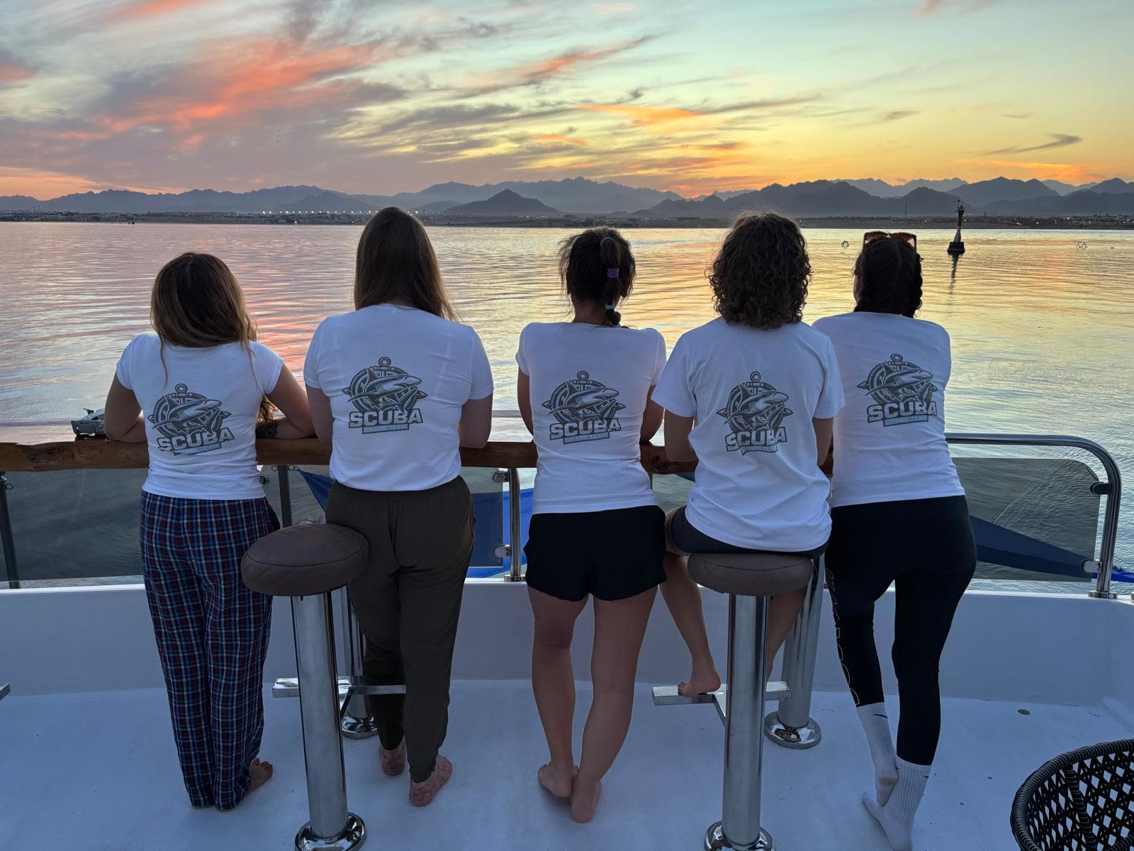Six people standing at the rail with their backs to the camera, watching the sun go down over the mountains.