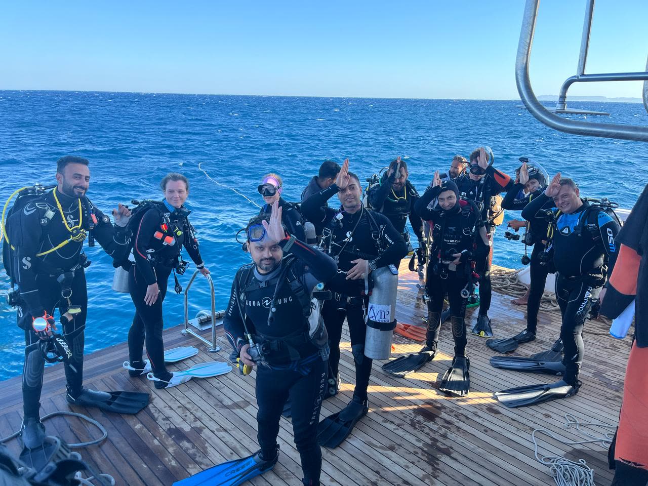 A dozen divers kitted up on the teak dive deck, fins laid out at their feet, flat blue water behind them.