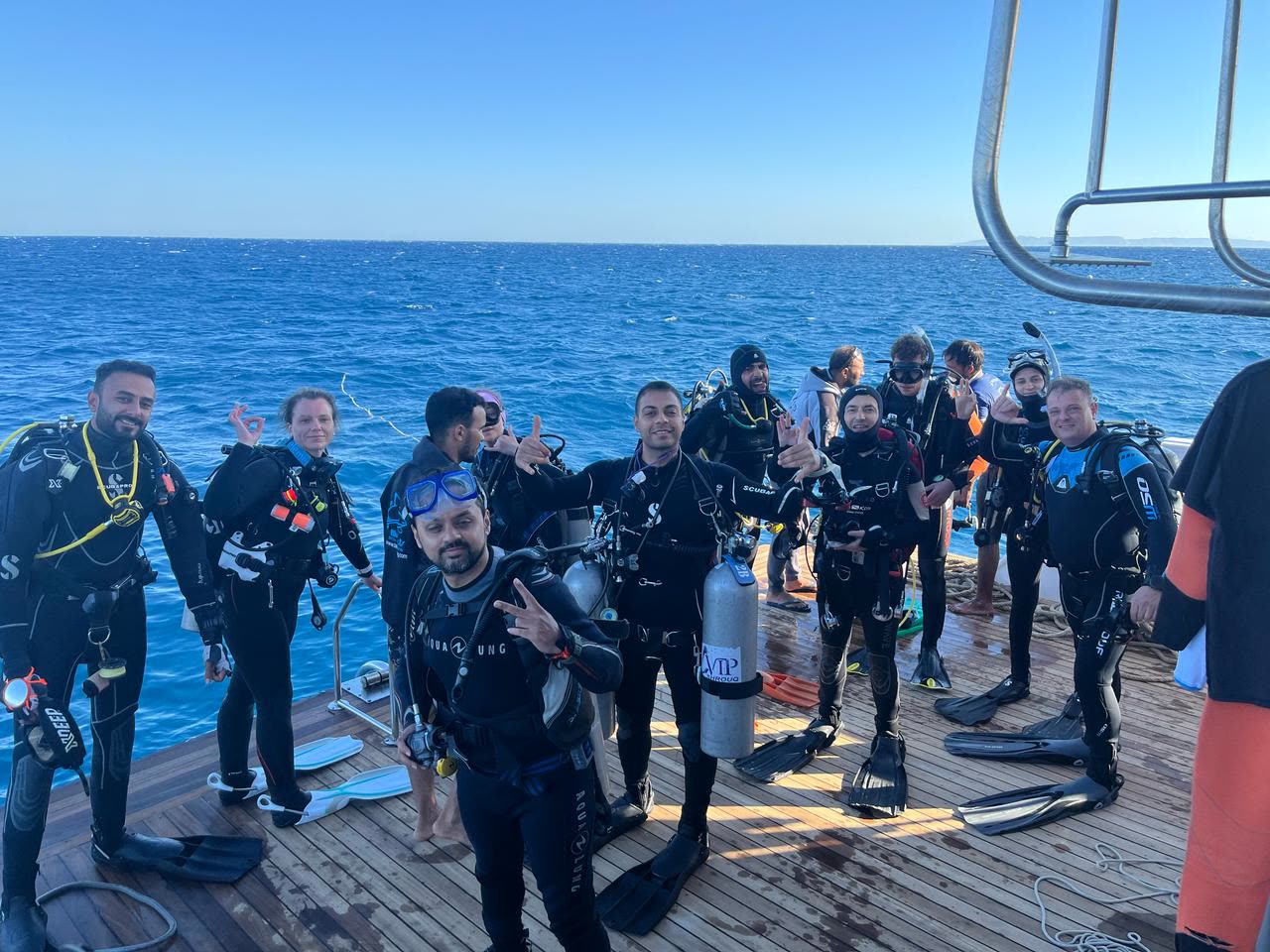 The full group on the dive deck in wetsuits and cylinders, waving, moments before the first entry of the day.