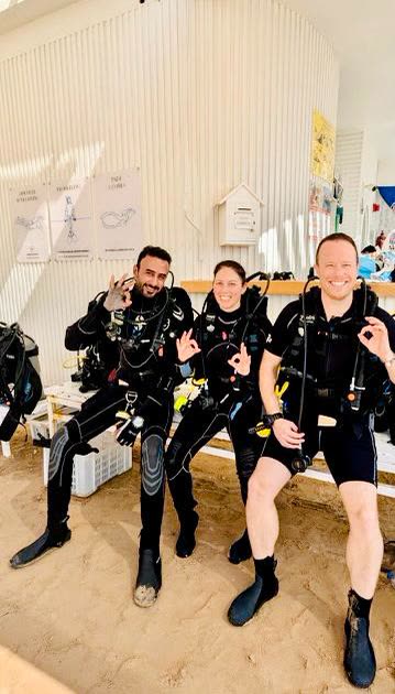 Three divers kitted up on a bench at the dive centre, all giving the OK sign.
