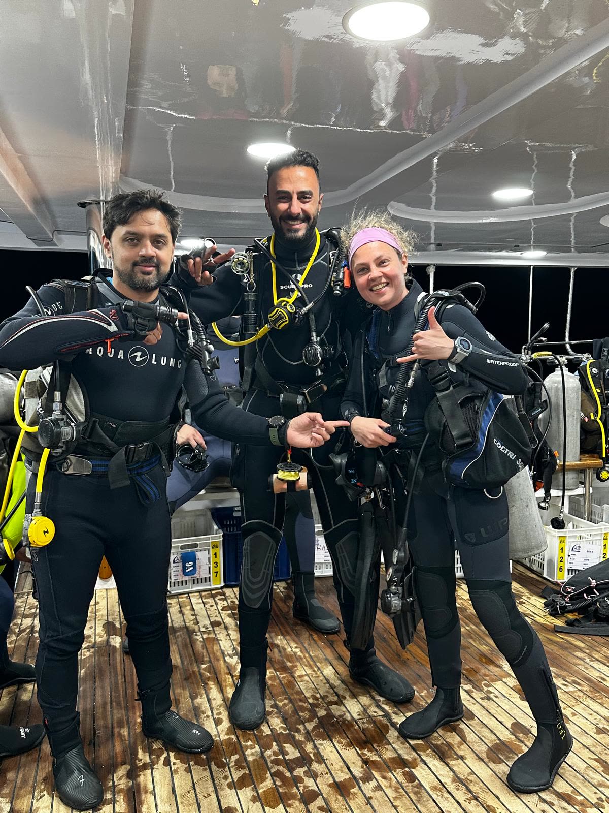 Three divers in full kit standing arm in arm on the dive deck, grinning, cylinders and regulators on their backs.