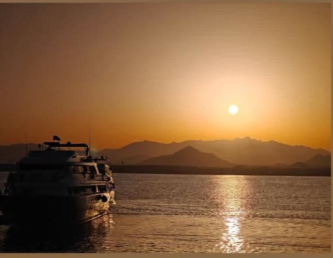 A boat silhouetted against the setting sun, mountains behind.